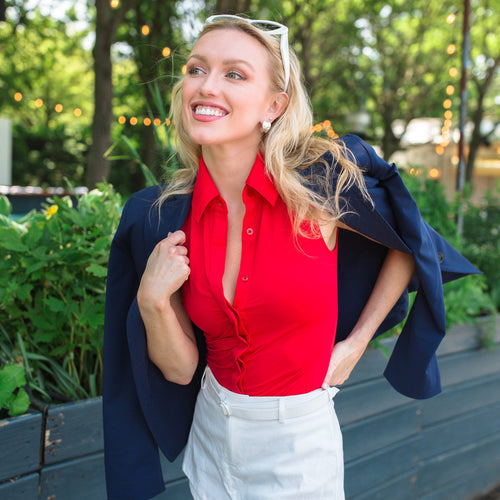 Woman in a red blouse and white shorts standing outdoors with greenery in the background