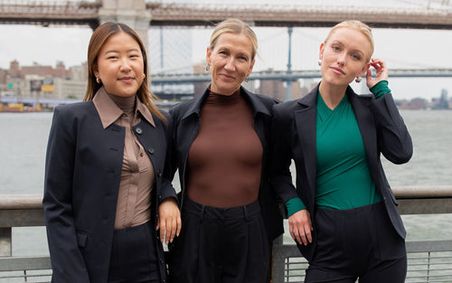 Three women standing together with a bridge and water in the background