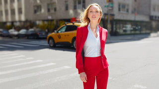 A woman in a red suit and light blue shirt walks across a city street with a yellow taxi and buildings in the blurred background.
