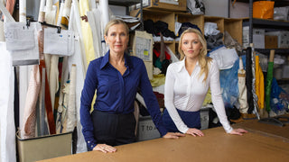 Two women standing behind a table in a storage or warehouse setting with shelves and boxes in the background.